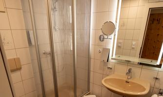 A modern bathroom with a glass shower and a sink. The walls are light-tiled, and a large mirror adds extra brightness.