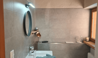 A modern bathroom with a cosmetic mirror and jewelry stand and a round mirror. The sink is equipped with a towel and a small decoration. The small window lets in a lot of light.