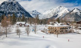 A picturesque winter landscape with snow-covered mountains in the background. In the foreground, there are some buildings and trees visible in the white blanket of snow.