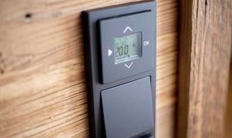 A modern thermostat with a digital display is mounted on a wooden wall. The temperature is set to 20 degrees.