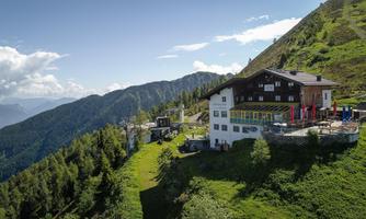 A picturesque mountain house surrounded by green hills and fir trees. In the background, majestic mountains and a blue sky can be seen.