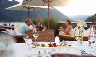A cozy restaurant terrace with a view of mountains and water. The table is festively set and there are guests sitting, enjoying drinks and conversing.