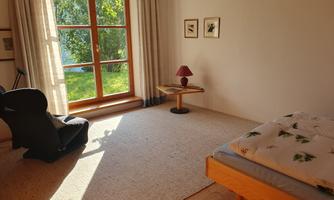 A cozy bedroom with a bed, a table, and a chair. Large windows let in plenty of sunlight and offer a view of greenery.