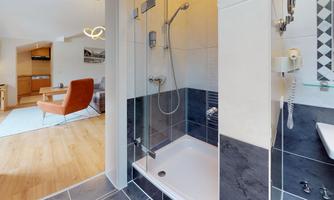 A modern bathroom with a shower and elegant tiles. In the background, there is a cozy area with an armchair and a table.
