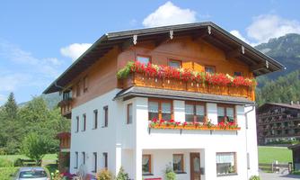A pretty house with wooden cladding and blooming flower balconies. In the background, green meadows and mountains are visible.