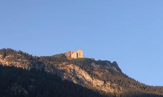 An impressive mountain landscape with steep rocks and a clear blue sky. The peak is surrounded by trees and shines in the sunlight.