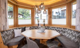 A cozy dining area with a round wooden table and upholstered benches. Large windows let in plenty of daylight and offer a view of the surroundings.