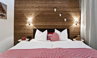 A cozy bedroom with a double bed and a wooden wall. The decoration includes cushions and small accessories in warm, rustic tones.