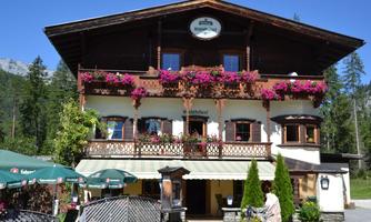 A traditional alpine hut with blooming flower balconies. The surroundings are surrounded by trees, and the architecture is rustic.