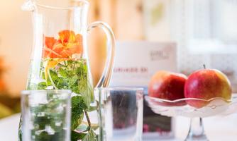 A glass carafe with fresh herbs and a flower is on the table. Next to it, two red apples are on a platter.