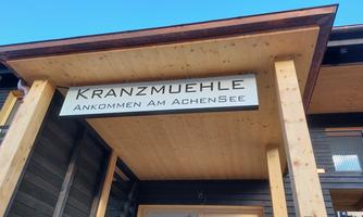 A wooden balcony with a sign that shows "Kranzmuehle" and "Arriving at Lake Achensee." The sky is blue and clear.
