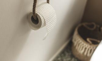A roll of toilet paper hangs from a rope on the wall. In the background, a toilet bowl and a basket are visible.
