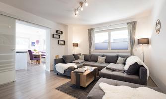 A modern living room with a gray sofa and a coffee table. The atmosphere is bright and inviting, with large windows and pleasant lighting.