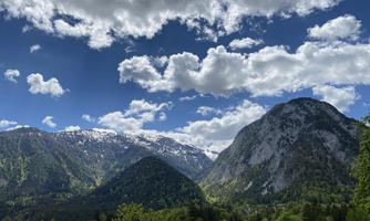 An impressive mountain landscape with snow-covered peaks and lush greenery. The sky is clear and shows many white clouds.