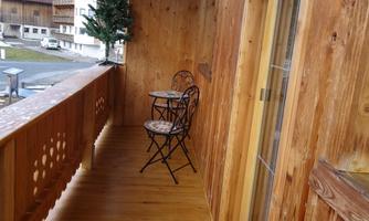 A cozy wooden balcony with a small table and two chairs. In the background, alpine buildings and a decorated tree are visible.