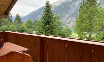 A terrace with a wooden railing and a view of a wooded mountain. In the background, green trees and a mountain landscape can be seen.