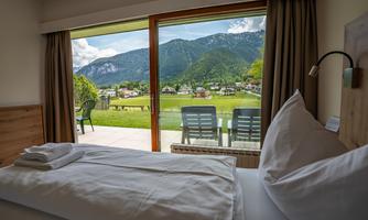 A cozy room with a view of the mountains. Outside, there is a terrace with seating options and lots of green nature.