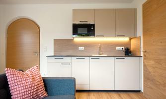 A modern kitchen unit with bright furniture and a microwave. A cozy living area with a gray sofa and a decorative pillow.