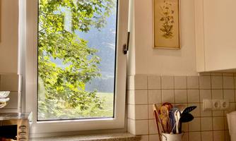 A modern kitchen with a window that overlooks a green garden view. On the countertop, there is a cutting board and kitchen utensils.