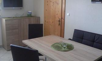 A modern dining area with a wooden table and black chairs. A television hangs on the wall and a wooden door leads outside.