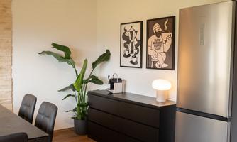 A modern kitchen with a black cabinet, a refrigerator, and a stylish lamp.  
Two artworks hang on the wall, and a plant brings life to the room.