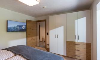 A modern bedroom with a large bed and a TV on the wall. The room is bright and minimally furnished with a wooden-look wardrobe.