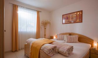 A cozy bedroom with a large bed and decorative pillows. The windows are fitted with light curtains and the room is brightly lit.