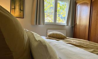 A cozy bedroom with a large window that lets in daylight and autumn colors. The room is decorated with a wood-colored wardrobe and soft pillows.