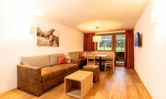 A modern living room with a cozy couch and a dining table. Large windows provide plenty of daylight and a view of the greenery.