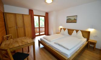 A bright room with a double bed, wooden furniture, and a table. There is a window overlooking a balcony.