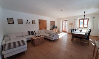A bright living room with a large, comfortable sofa and a wooden table. Large windows let in plenty of light and the decor is simple and cozy.