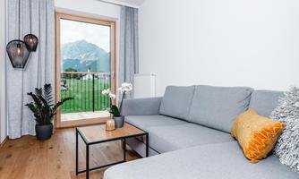 A modern living room with a gray sofa and a wooden table.  
Windows with a view of the mountains and a beautiful plant arrangement.