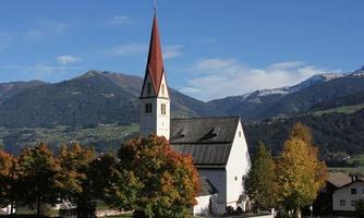 A beautiful church with a steeple stands in front of a picturesque mountain landscape. Colorful trees surround the building and the sky is clear and blue.