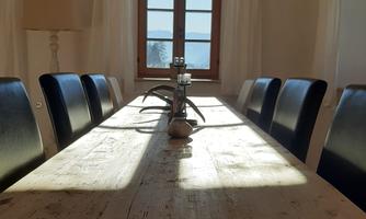 A large wooden dining table with black chairs is located in a bright room. Sunlight shines through the window and offers a view outside.