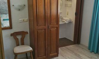 A simple room with a wooden wardrobe, a chair, and a colorful carpet. In the background, a bathroom can be seen.