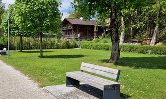 A wooden bench stands on a green meadow surrounded by trees. In the background, a rustic building is visible.