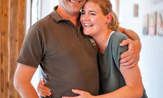 A laughing couple stands in a bright hallway. They embrace each other and appear happy.