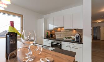 A modern kitchen with white cabinets and wooden details. In the foreground, there is a wine bottle and a glass on the counter.