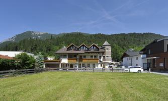 A modern house in the midst of a green meadow, surrounded by mountains and a clear sky. In the background, trees and additional buildings are visible.