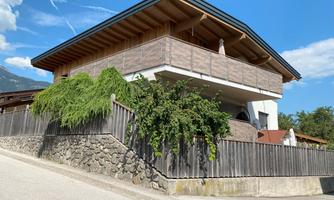 A modern house with wooden cladding and a spacious balcony. It is surrounded by a garden with green plants.