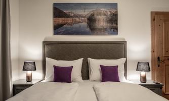 A cozy bedroom with a double bed and pillows in purple. Pictures hang on the walls, and two bedside lamps provide soft light.