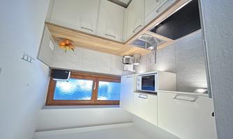 A modern kitchen with white furniture and a wooden countertop. There is a privacy screen above the window and fruit in a bowl.