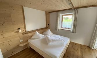 Unified bedroom with light wood and a large window. The room is minimally furnished, featuring a double bed and two pillows.