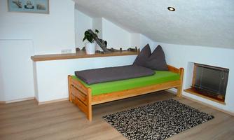 A cozy room under the roof with a wooden bed and green lounging area. A small rug and plants create an inviting atmosphere.