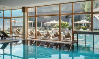 An elegant indoor pool with large windows and a view of a sunny terrace. Loungers are arranged around the pool to provide relaxation.