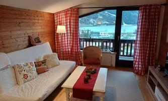 A cozy living room with a white sofa and checked curtains. The large window offers a beautiful view of the mountains and the water.