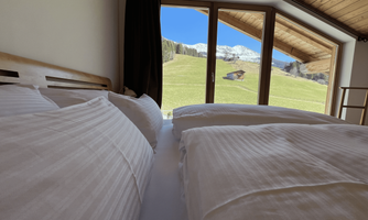 A cozy bedroom with two beds and a view of green meadows and mountains. Large windows let in plenty of light.