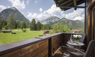 A balcony with a table and chairs, surrounded by a stunning mountain landscape. In the background, green meadows and tall mountains can be seen under a blue sky with clouds.