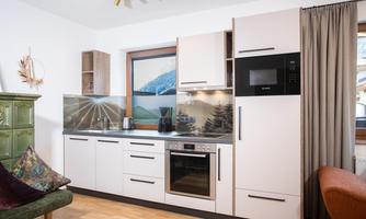 A modern kitchen with light furniture and a window view of a mountain landscape. The room is well-lit and stylishly furnished.