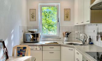 A bright, modern kitchen with a window that looks out onto a green view. It is equipped with modern appliances and a small dining table.